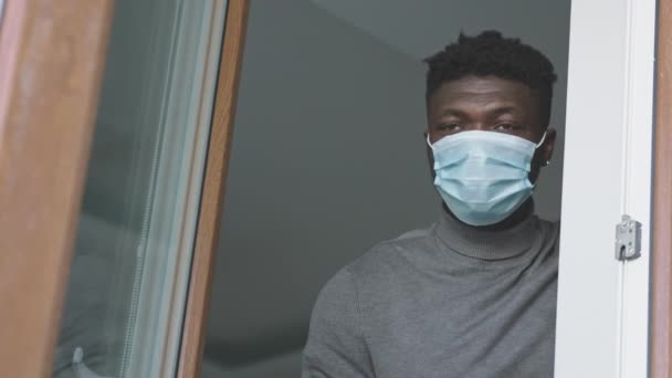 Afrikanischer schwarzer Mann mit medizinischer Maske steht am Fenster und schaut nach draußen. Quarantäne und Konzept für psychische Gesundheit — Stockvideo