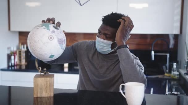 Samotny czarny podróżnik patrzący na globus w swoim domu w masce medycznej. Podróżowanie podczas pandemii zakrzepowo-19 — Wideo stockowe