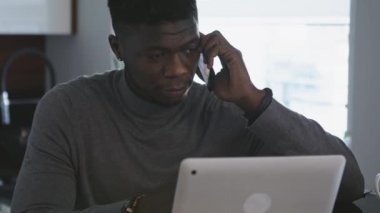 Afrikalı Amerikalı siyahi iş adamı bilgisayarına bakarken dairesinde telefon görüşmesi yapıyor.