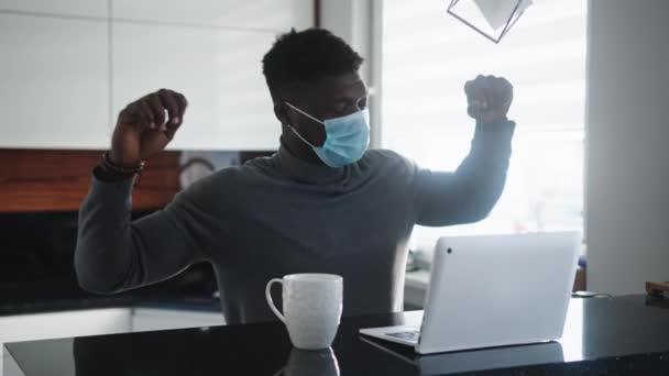 Молодой афроамериканец черный мужчина растягивается во время работы над ноутбуком в своей квартире с маской на лице — стоковое видео