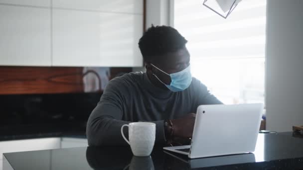 Молодой афроамериканец черный мужчина, работающий над ноутбуком в своей квартире в маске для лица — стоковое видео