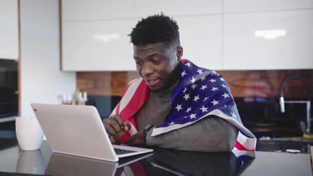 Africký Američan s vlajkou USA přes ramena sleduje hru na svém notebooku s šálkem čaje na stole. Překvapený výraz obložení — Stock video