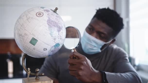 Ung svart man med ansiktsmask tittar på genom förstoringsglas. Reseförbud under en global pandemi — Stockvideo