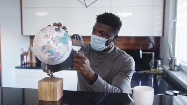 Joven hombre negro con mascarilla mirando la lupa a través. Prohibición de viajar durante la pandemia mundial — Vídeo de stock