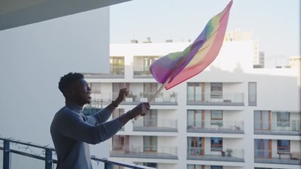 Счастливый молодой черный мужчина размахивает радужным флагом с балкона — стоковое видео