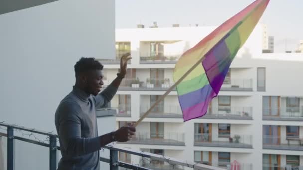Glücklicher junger schwarzer Mann schwenkt Regenbogenfahne vom Balkon — Stockvideo
