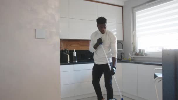 Bonito homem negro dançando e cantando enquanto limpa o chão na cozinha. conceito de vida doméstica — Vídeo de Stock
