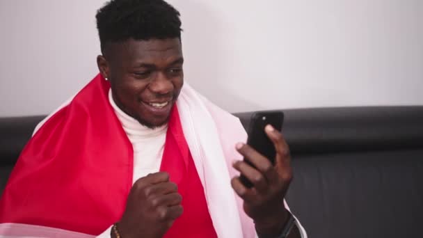 Молодий щасливий чорний чоловік з польським прапором дивиться гру, що транслюється на смартфоні — стокове відео