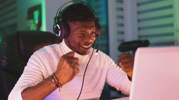 Feliz joven negro con auriculares iluminados usando el ordenador portátil. Oficina en el hogar y concepto de trabajo remoto. Retrato del comerciante de acciones — Foto de Stock