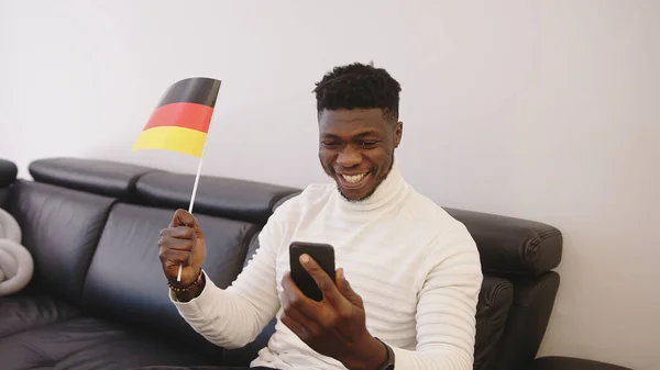 Genç siyahi adam akıllı telefondan maç izleyip Alman bayrağı sallıyor. — Stok fotoğraf