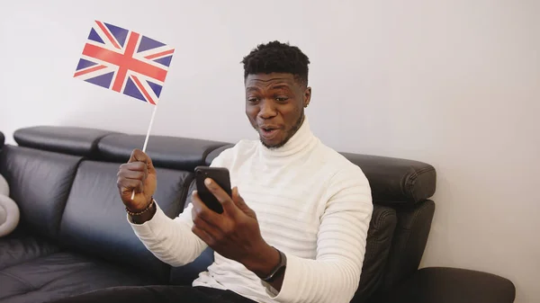 Genç siyahi adam akıllı telefondan maç izleyip İngiliz bayrağı sallıyor. — Stok fotoğraf