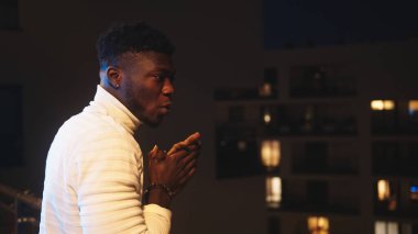 Genç siyah adam soğuk bir gecede balkonda duruyor ve ellerini ovuşturuyor.