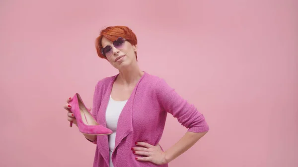 Mulher madura com cabelo curto vermelho segurando sapatos curativos altos Isolado no fundo roxo — Fotografia de Stock