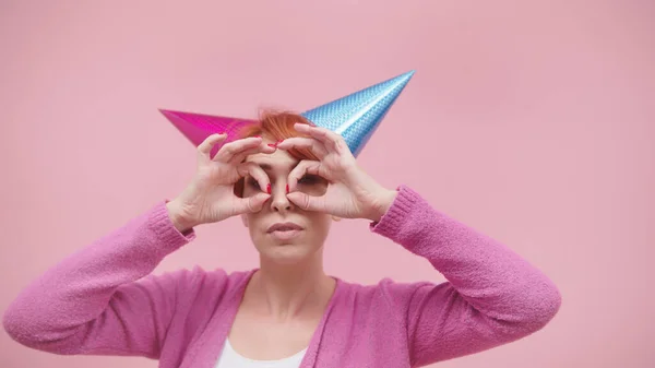 Verrückte Frau mittleren Alters mit 2 Party-Hüten und Fingern, die eine Brille über ihren Augen auf rosa Hintergrund bilden — Stockfoto