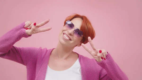 Schöne Frau in den Vierzigern mit einer rosafarbenen herzförmigen Brille, die mit beiden Händen ein Friedenszeichen zeigt. Vereinzelt auf rosa lila Hintergrund — Stockfoto