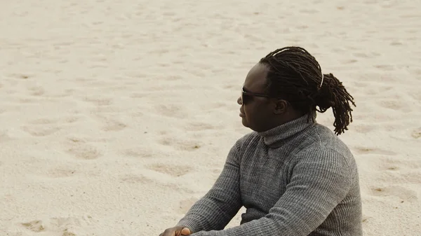 Jovem negro afro-americano sentado na areia em suéter e relaxante nas férias de inverno — Fotografia de Stock