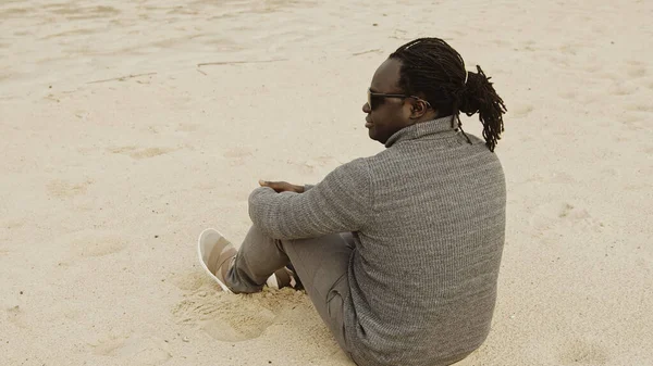 Mladý africký muž pocit chladu, zatímco sedí ve svetru na písečné pláži — Stock fotografie
