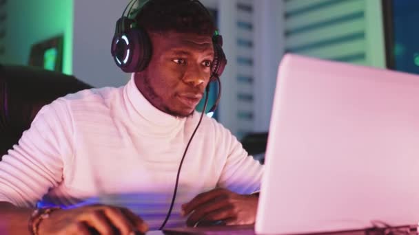 Felkent fiatal fekete férfi megvilágított fejhallgatóval, laptoppal. Otthoni iroda és távmunka koncepció — Stock videók