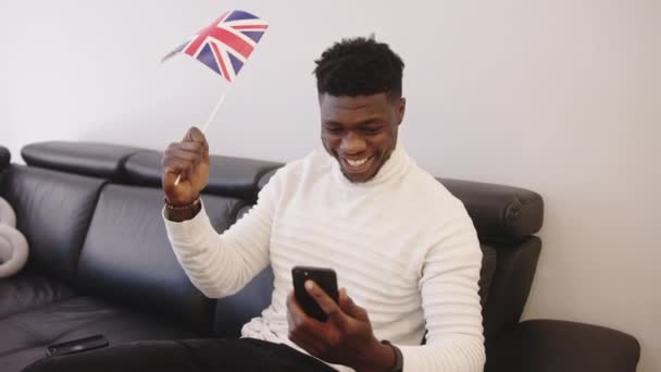 Genç siyahi adam akıllı telefondan maç izleyip İngiliz bayrağı sallıyor. — Stok video