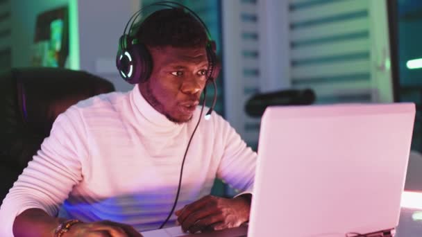 Csalódott fiatal fekete férfi megvilágított fejhallgatóval, laptoppal. Otthoni iroda és távmunka koncepció — Stock videók