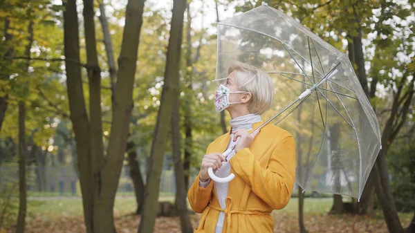 Schöne blonde Frau mit Gesichtsmaske, die im Herbst im Park einen durchsichtigen Regenschirm dreht. Coronavirus-Ausbruch 2020 — Stockfoto