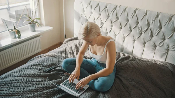 Mujer joven usando portátil en su dormitorio. Freelancer trabajando desde la comodidad de su hogar — Foto de Stock