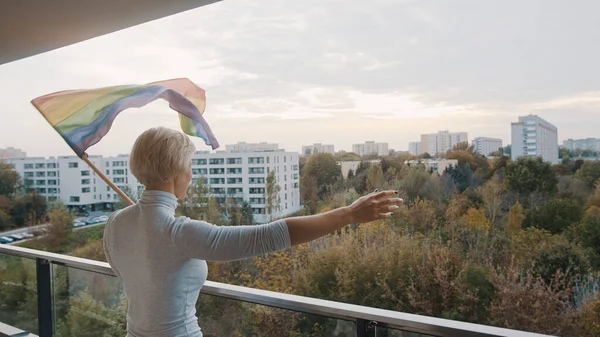 Jonge blonde blanke vrouw zwaait met regenboogvlag vanaf het balkon. Steun voor LGBTQ — Stockfoto