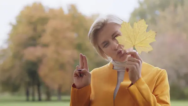 Junge blonde Frau mit gelbem Herbstblatt und Friedenszeichen mit den Fingern — Stockfoto