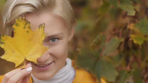 Portraitaufnahme einer jungen kaukasischen blonden Frau mit blauen Augen, die ihr Gesicht mit gelbem Herbstblatt verdeckt — Stockfoto