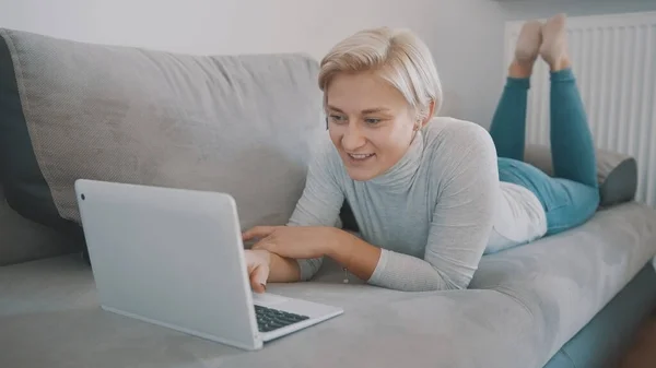 Mujer joven charlando con amigos en las redes sociales utilizando el ordenador portátil en su sofá — Foto de Stock
