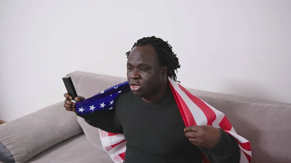 African american black man with usa flag over the shoulders watching sport game on tv on his couch
