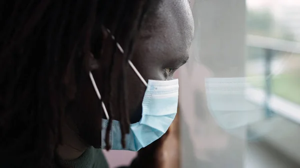Тривожний чорношкірий афроамериканський чоловік з медичною маскою дивиться крізь вікно — стокове фото