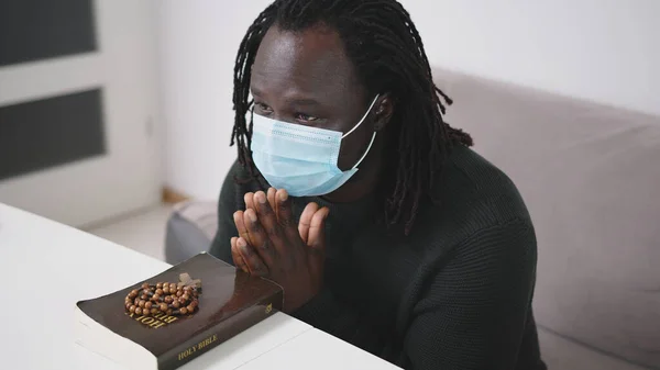 Yüz maskeli siyah Afrikalı adam kutsal kitap ve tespih duası yapıyor. Covid-19 Virüs Salgını sırasında Dinde Umut Aranıyor — Stok fotoğraf