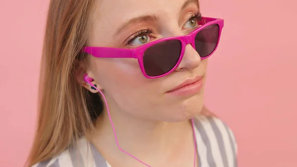 Молодая белая женщина в солнечных очках слушает музыку в наушниках. Изолированный на розовом фоне, крупным планом — стоковое фото