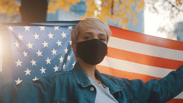 USA presidential election concept. Proud woman with face mask proudly holding american flag outdoors