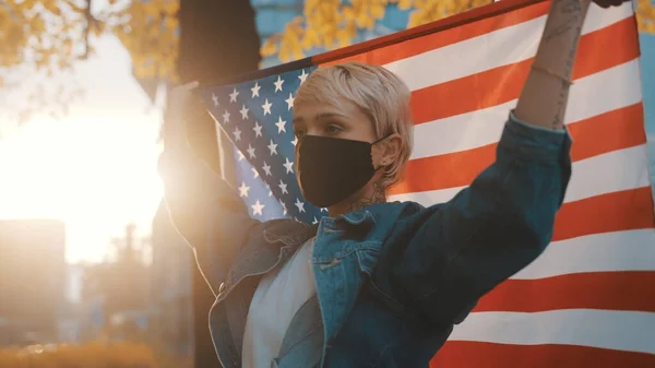 Mujer joven con máscara facial sosteniendo orgullosamente la bandera americana al aire libre.Concepto de las elecciones presidenciales de Estados Unidos. — Foto de Stock