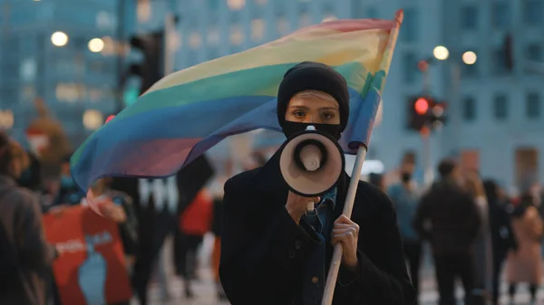 Mujer joven con máscara facial hablando en el megáfono mientras sostiene la bandera del arco iris en la multitud —  Fotos de Stock