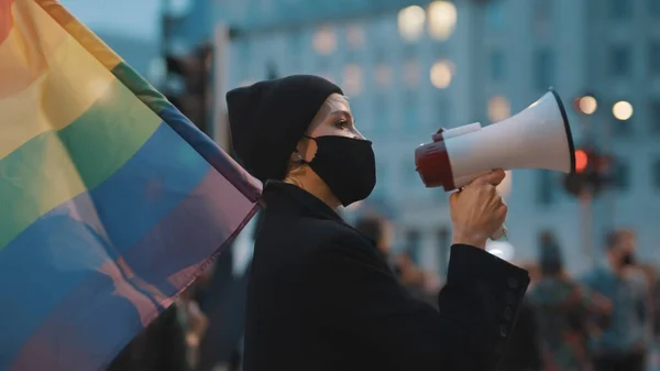 Jovem com máscara facial falando no megafone enquanto segurava a bandeira do arco-íris na multidão — Fotografia de Stock