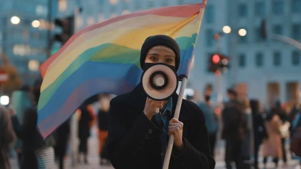 Mujer joven con máscara facial hablando en el megáfono mientras sostiene la bandera del arco iris en la multitud —  Fotos de Stock