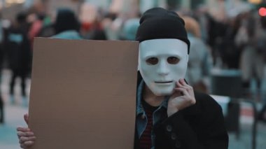 Genç bir kadın kalabalıkta elinde siyah karton tutarken tiyatro maskesini çıkarıyor. Ayrımcılığa karşı protesto