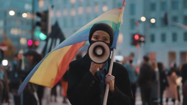 Νεαρή γυναίκα με μάσκα προσώπου που μιλάει στο μεγάφωνο κρατώντας τη σημαία του ουράνιου τόξου μέσα στο πλήθος — Αρχείο Βίντεο