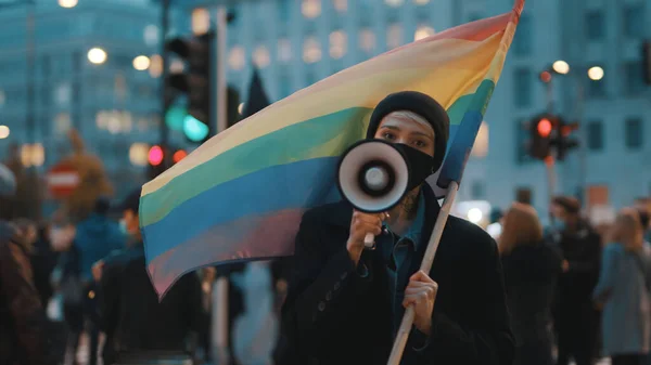 Jonge vrouw met gezichtsmasker spreken in de megafoon terwijl het houden van regenboog vlag in menigte — Stockfoto