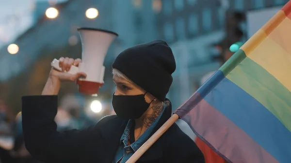 Mujer joven con máscara facial y megáfono sosteniendo la bandera del arco iris en multitud — Foto de Stock