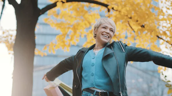 Jovem mulher caucasiana feliz em jaqueta de couro com as mãos estendidas girando sob a árvore no parque no dia de outono — Fotografia de Stock