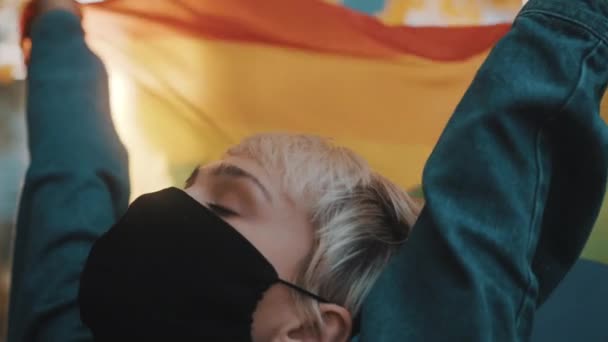 Stolt ung blond kvinna med ansiktsmask viftande regnbåge flagga. Närbild — Stockvideo