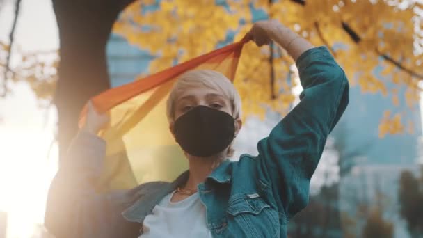 Orgogliosa giovane donna bionda con maschera viso sventola bandiera arcobaleno sotto l'albero con foglie gialle — Video Stock