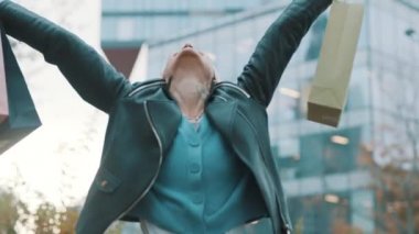 Alışveriş poşetleriyle mutlu genç bir kadın alışveriş merkezinin önünde heyecanla gülüyor.