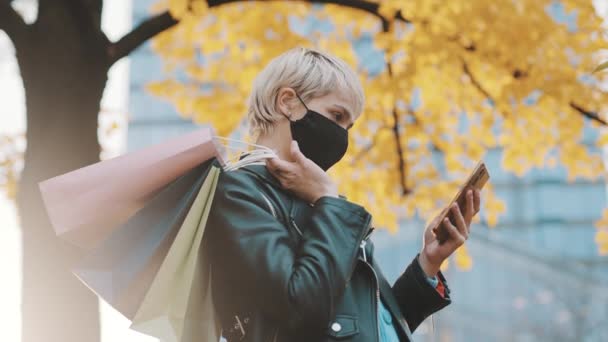 Portret van een jonge vrouw in leren jas en papieren boodschappentassen over de schouder met smartphone — Stockvideo