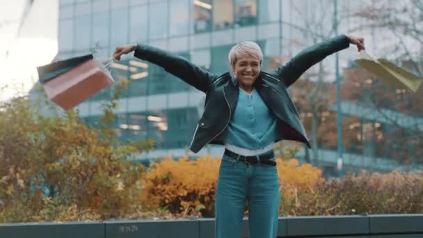 Glückliche junge Frau mit Einkaufstüten, die lachend und aufgeregt vor dem Einkaufszentrum hüpft — Stockvideo