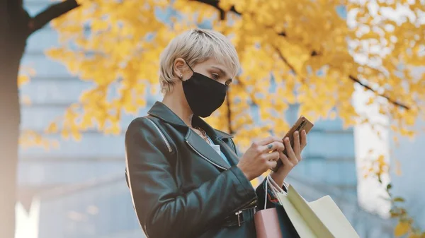 Jovem com máscara facial e sacos de compras usando smartphone sob a árvore com folhas amarelas — Fotografia de Stock